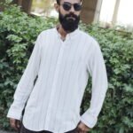 Man wearing a white premium textured striped shirt with sunglasses, standing outdoors near greenery and a sidewalk, styled for a modern yet traditional look.
